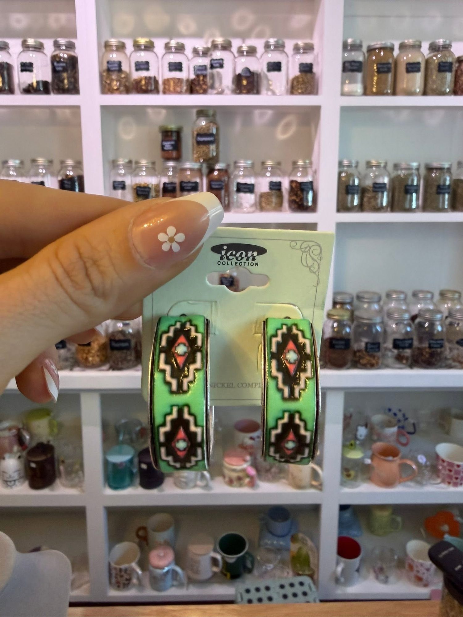 Hand holding a pair of green Southwest-pattern hoop earrings in front of a shelf of mini display mugs